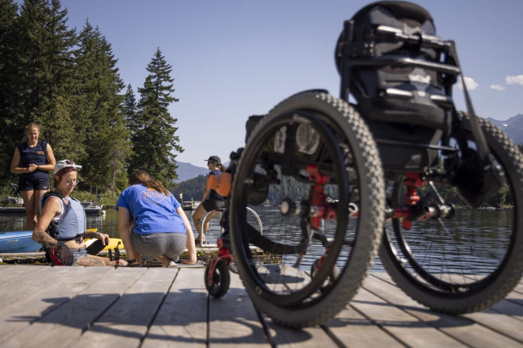 A group of people assist a woman in a kayak on a lakeside dock, with a wheelchair prominently placed in the foreground. The scene is surrounded by forested mountains under a clear blue sky, reflecting inclusive outdoor recreation.