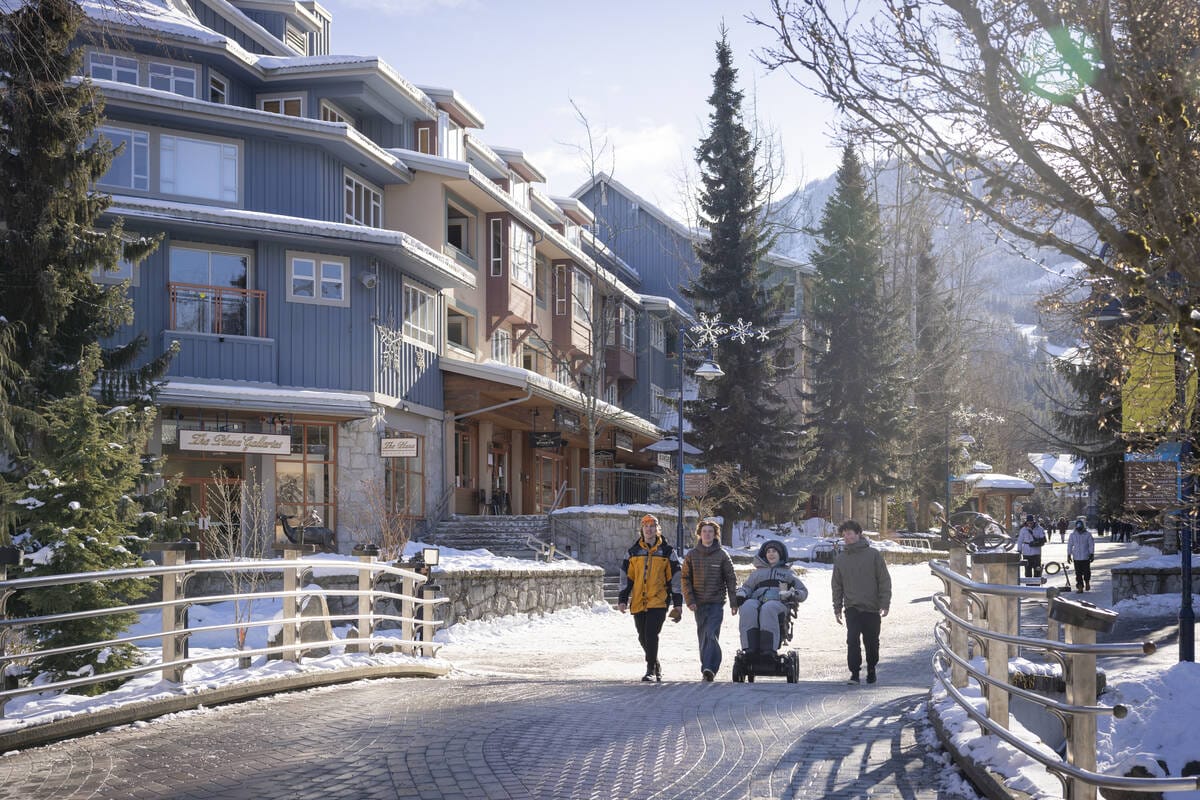 group of friends, including one in a wheelchair, enjoys a sunny winter day in Whistler Village, with snow-covered mountains and cozy accommodations in the background.
