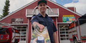 Firefighter in a navy uniform extends an evacuation brochure toward the camera outside a red fire station with trucks nearby.