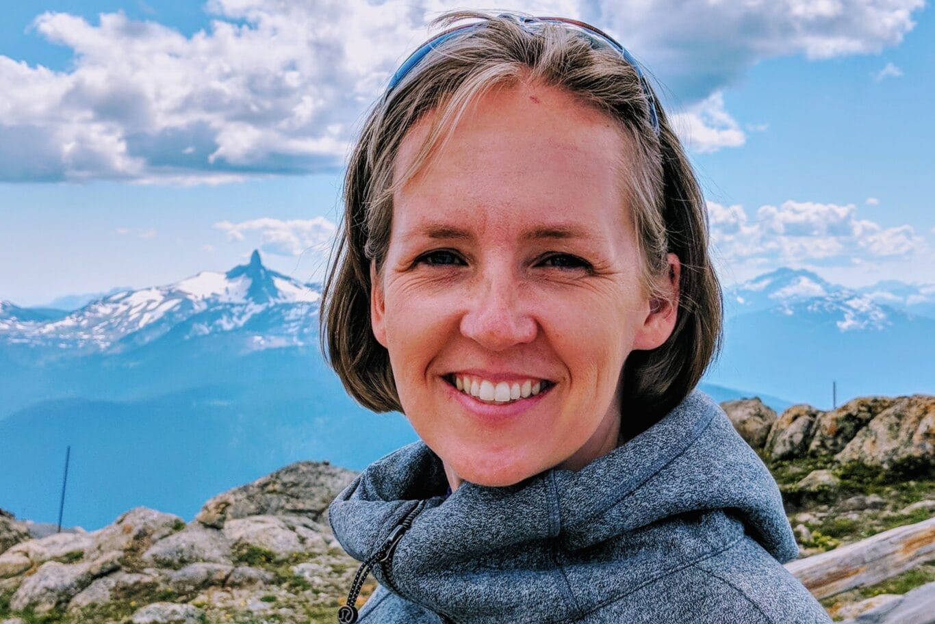 Woman smiling at the camera on a rocky mountaintop with snow-capped peaks in the background, under a blue sky.