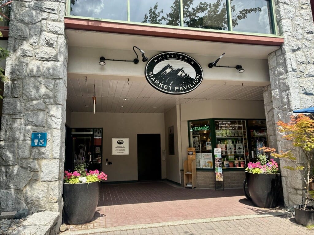 Entrance to Whistler's Market Pavilion with stone pillars, circular sign, and flower planters outside a shop storefront