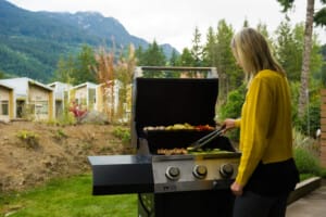 Woman in a yellow sweater tending food on a black outdoor grill in a backyard with mountains and trees in the background.