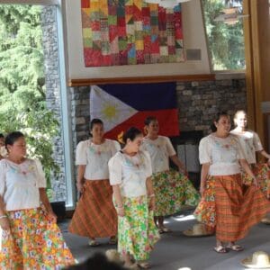 Group of Filipino women in white blouses and colorful skirts performing a traditional dance on stage, with a Philippine flag in the background.