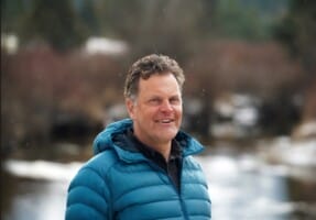 Smiling man in a teal-blue puffer jacket outdoors on a snowy winter day.