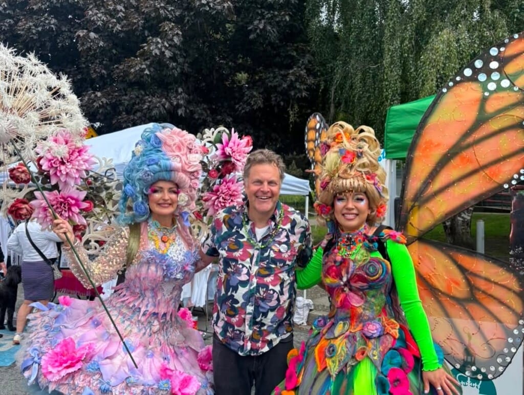 Three people in colorful costumes pose at an outdoor festival, two women in elaborate floral outfits and a man in a tropical shirt, with butterfly wings visible on the right.