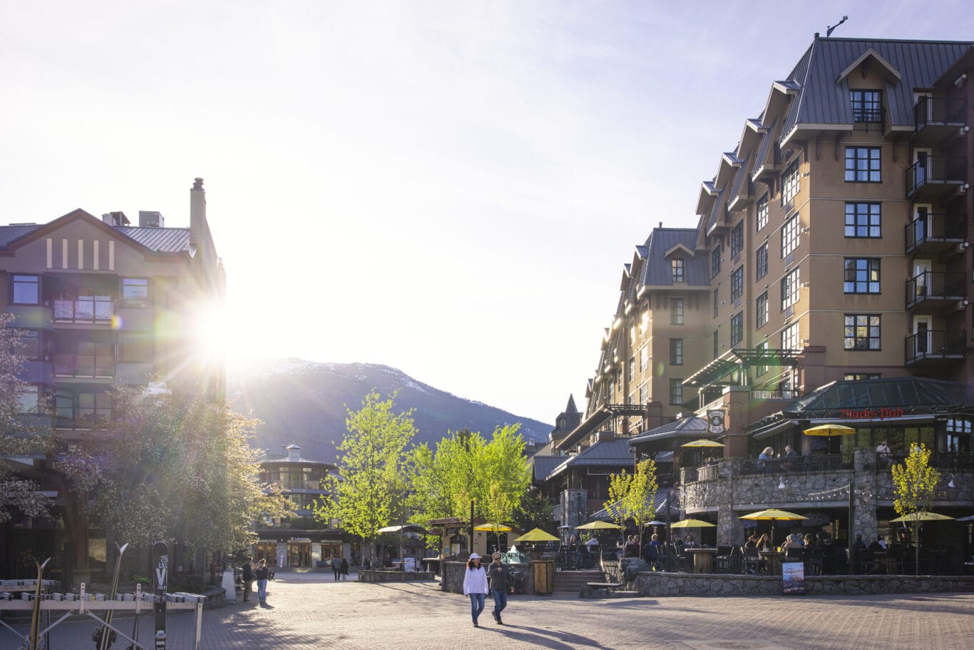Sunlit town square with European-style buildings, outdoor cafe seating under yellow umbrellas, and pedestrians.