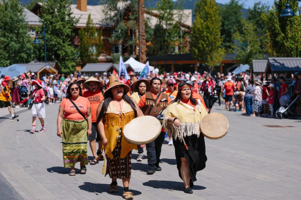 Group of Indigenous people in traditional dress marching in a street parade, some drumming, with spectators and tents in the background.