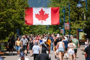 Crowd of people walking along a sunny street beneath a large Canadian flag overhead, creating a festive atmosphere.