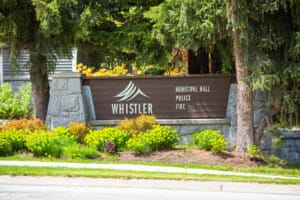 Whistler municipal sign reading Municipal Hall, Police, Fire with a mountain logo, set in a stone planter amid yellow flowers and evergreens.